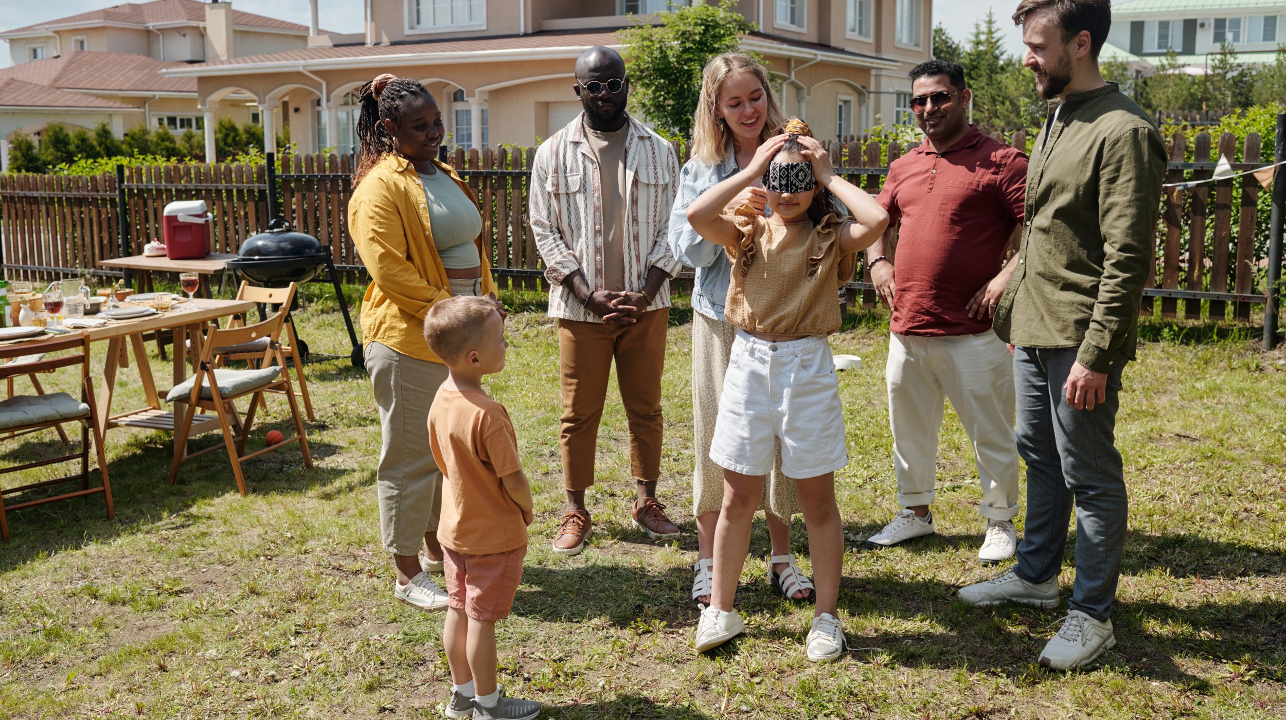 Young woman blindfolding little daughter while playing hide and seek with friends on green grass of backyard of country house